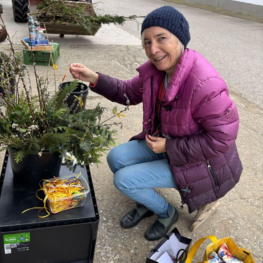 Eine Frau kniet neben einer großen Topfpflanze, hält einen Zweig und lächelt für ein Foto.