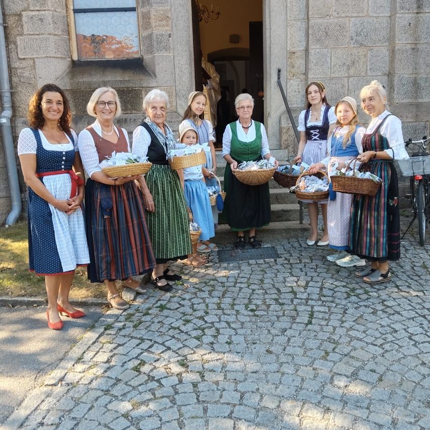 A group of women and children dressed in traditional clothing stand outside a stone building, holding baskets. A bicycle is parked nearby.