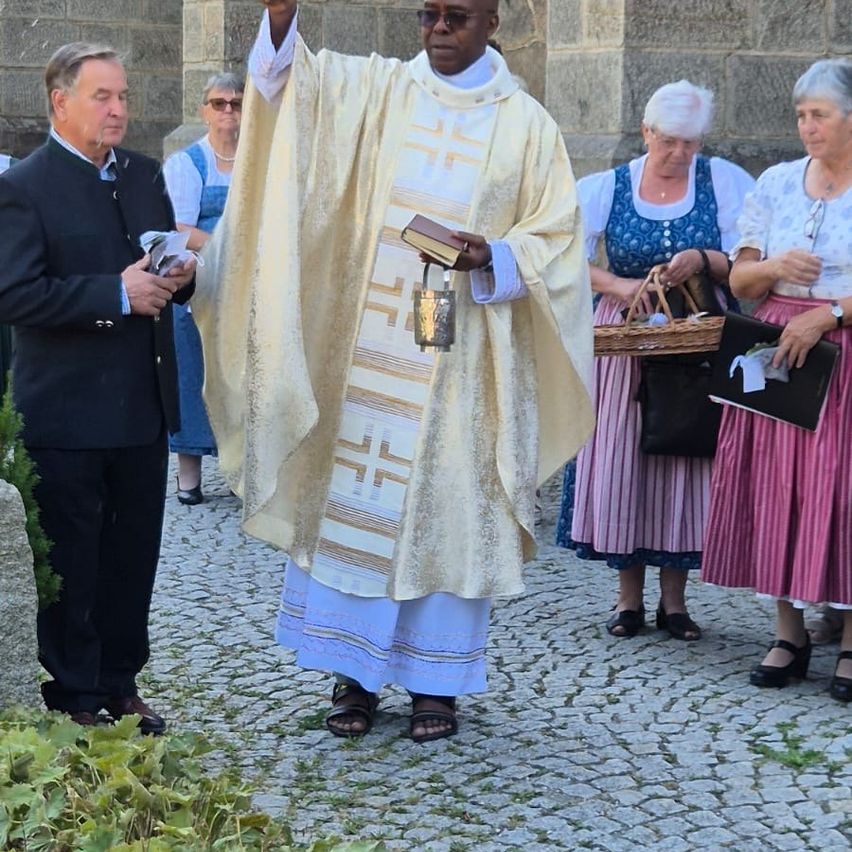 A priest in a white and gold robe holds a book and a bell, standing in front of a group of people, two of whom are holding baskets.