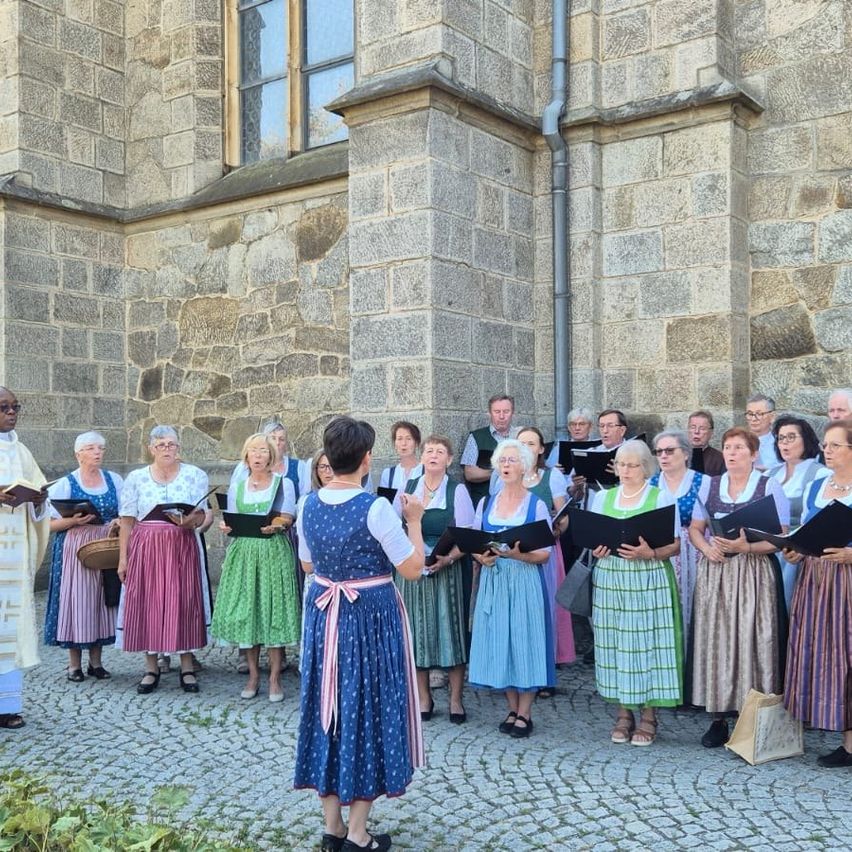 A choir of women in traditional attire singing in front of a stone building.