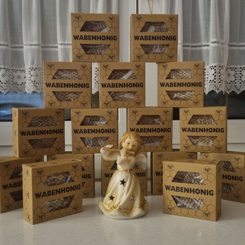 Ein Stapel Kartonboxen mit dem Wort 'Wabenhonig' darauf wird vor einem weißen Vorhang ausgestellt. Eine Engelsfigur steht in der Mitte.