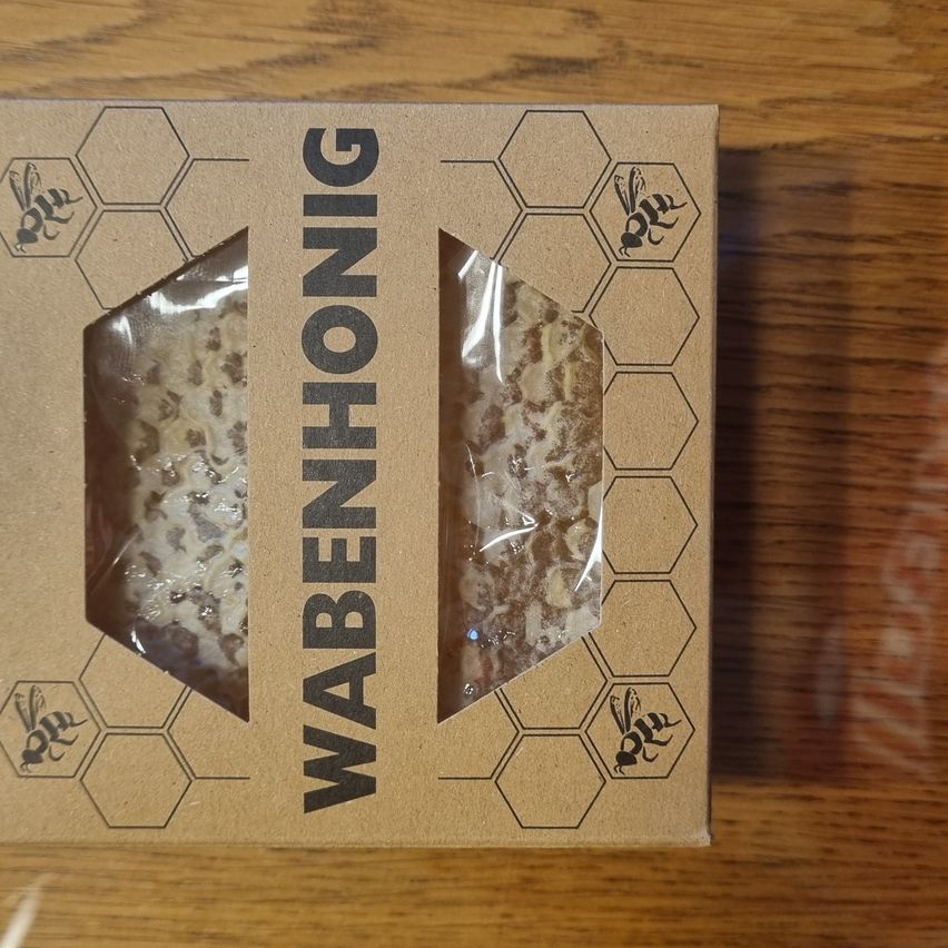 Eine Pappschachtel zeigt ein Produkt namens Wabenhonig, mit einem Wabenmuster und einer Biene auf der Seite. Das Produkt scheint Honig mit einer Wabenstruktur zu sein, der mit Plastik bedeckt ist.