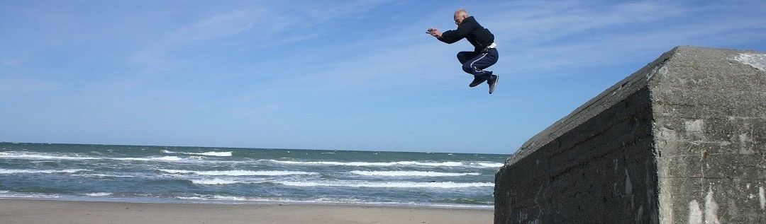 Ein älterer Mann in einem schwarzen Outfit springt über eine Betonbarriere in der Nähe des Ozeans. Der Himmel ist blau mit einigen Wolken, und das Meer hat kleine Wellen.