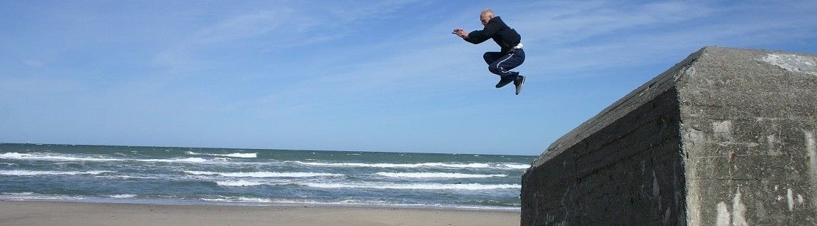 Ein älterer Mann in einem schwarzen Outfit springt über eine Betonbarriere in der Nähe des Ozeans. Der Himmel ist blau mit einigen Wolken, und das Meer hat kleine Wellen.
