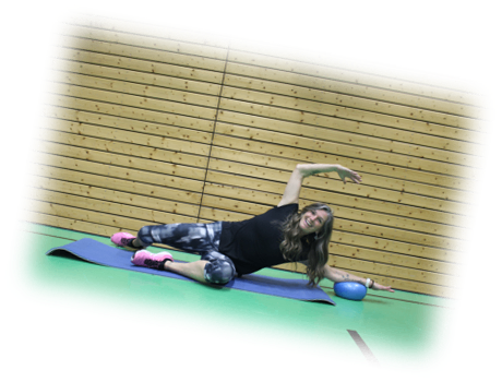 Eine Frau in einem schwarzen Shirt macht Yoga auf einer Matte. Sie hat eine blaue Kugel und lächelt. Sie befindet sich in einem Fitnessstudio mit einer Holzwand.