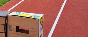 Ein Athlet springt über eine Hürde auf der Laufbahn. Im Vordergrund sind Pappkartons gestapelt.