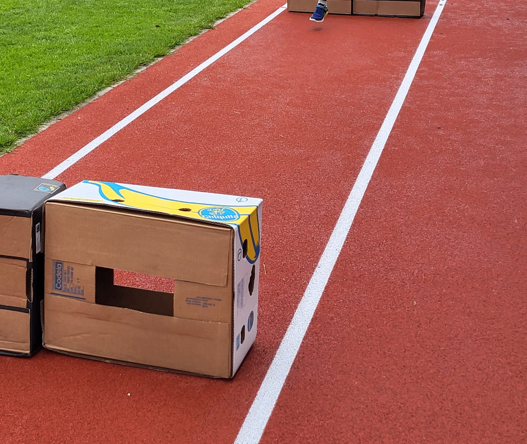 Ein Athlet springt über eine Hürde auf der Laufbahn. Im Vordergrund sind Pappkartons gestapelt.
