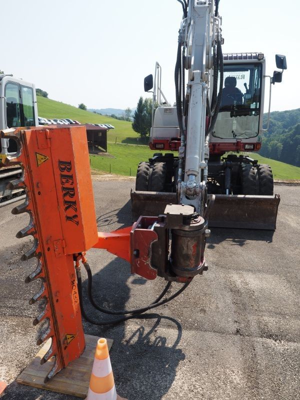 Ein orangefarbenes Maschinenfahrzeug mit dem Wort 'BERKY' ist auf der Straße geparkt. Eine Person sitzt in der Kabine. Vor dem Fahrzeug steht ein Verkehrskegel.