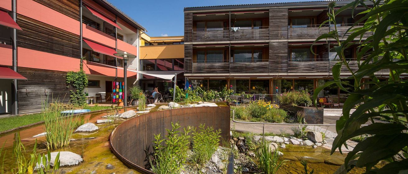 Ein Blick auf das Äußere eines Gebäudes mit Garten, Teich und Brunnen. Einige Personen sitzen auf Stühlen vor dem Gebäude.