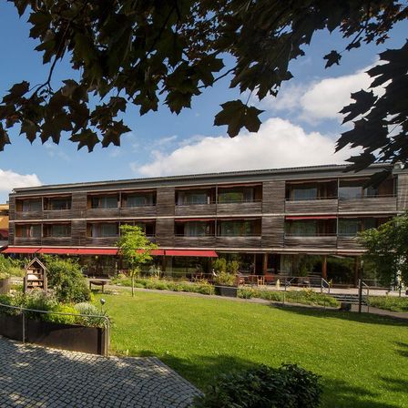 Ein Weg in einem grünen Gartenbereich zwischen zwei modernen Gebäuden mit roten Markisen und Balkonen. Der Garten hat kleine Pflanzen, ein Vogelhaus und einen Baum.