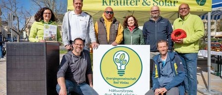 Eine Gruppe von Menschen steht unter einem Zelt mit der Aufschrift 'Kräuter & Solar' und lächelt für ein Foto. Ein Schild mit einer Glühbirne und den Worten 'Energiegemeinschaft Bad Vöslau' ist vorhanden.