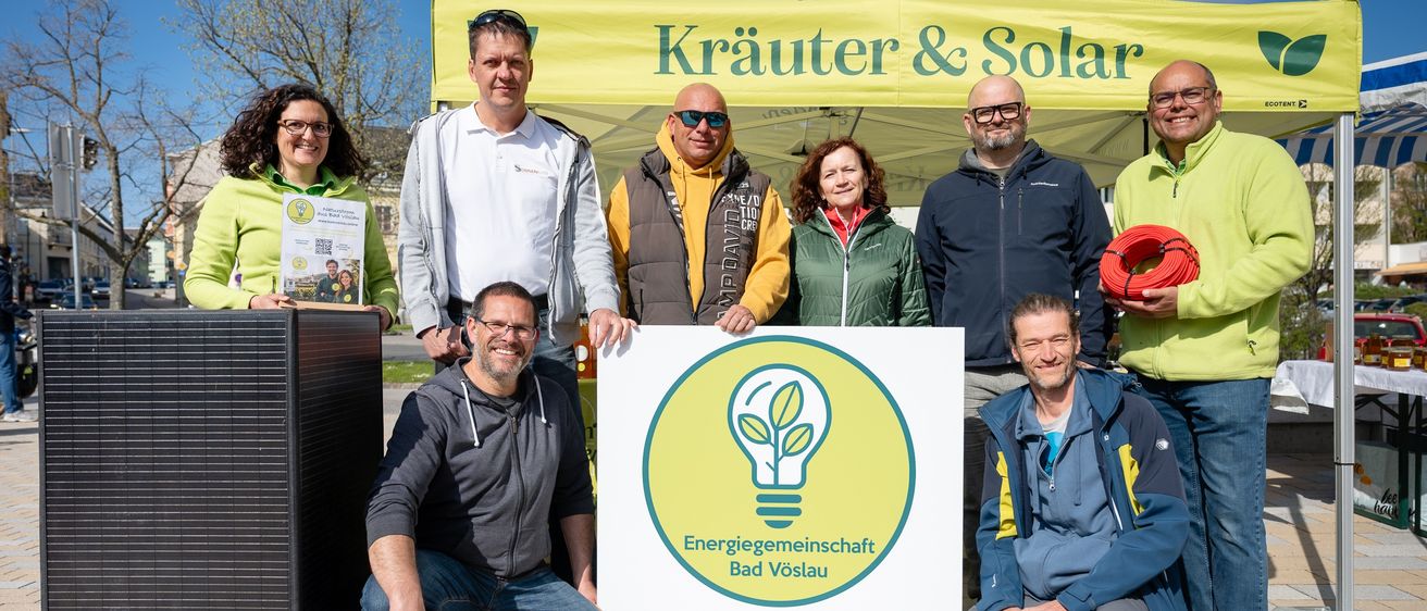 Eine Gruppe von Personen posiert für ein Foto unter einem gelben Zelt mit der Aufschrift 'Kräuter & Solar' und hält ein Schild für 'Energiegemeinschaft Bad Vöslau', ein Naturstromprojekt.