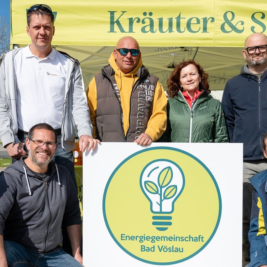 Eine Gruppe von Personen posiert für ein Foto unter einem gelben Zelt mit der Aufschrift 'Kräuter & Solar' und hält ein Schild für 'Energiegemeinschaft Bad Vöslau', ein Naturstromprojekt.
