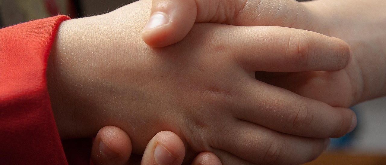 Zwei Hände von Kindern, die ineinander verschränkt sind, zeigen Daumen, Zeige-, Mittel-, Ring- und Kleinfinger. Die Hand des Kindes links trägt ein rotes Langarmshirt.
