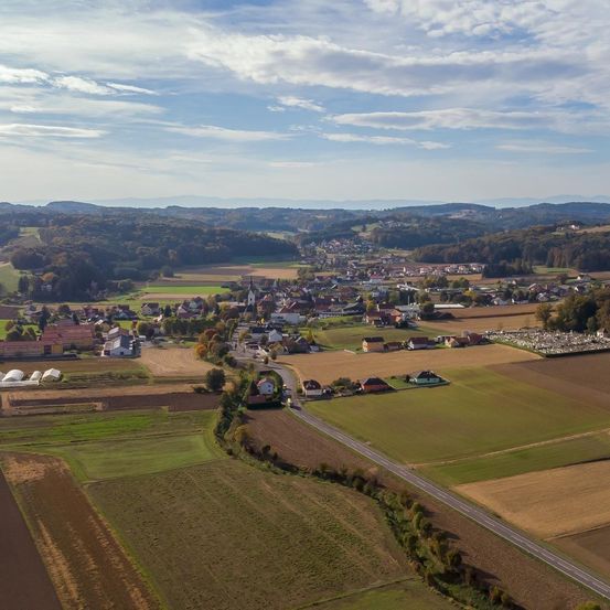 Luftaufnahme eines kleinen Dorfes umgeben von Feldern und Bergen unter einem blauen Himmel mit verstreuten Wolken.