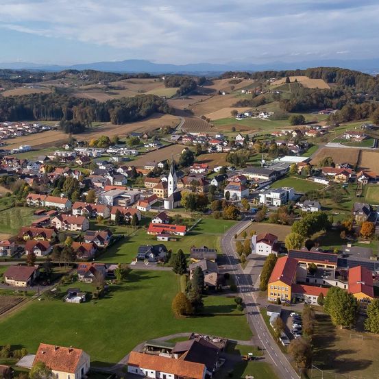 Luftaufnahme einer kleinen Stadt mit vielen Häusern, einige mit roten Dächern, grünen Feldern und einem klaren Himmel.