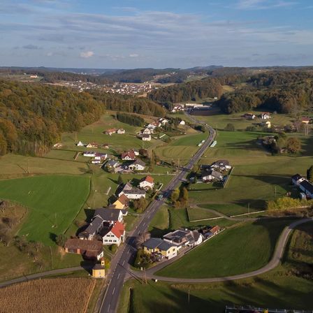 Luftaufnahme eines ländlichen Gebiets mit grünen Feldern und einer kurvenreichen Straße, umgeben von Häusern und Bäumen, unter einem klaren blauen Himmel mit einigen Wolken.