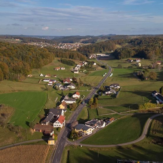 Luftaufnahme eines ländlichen Gebiets mit grünen Feldern und einer kurvenreichen Straße, umgeben von Häusern und Bäumen, unter einem klaren blauen Himmel mit einigen Wolken.