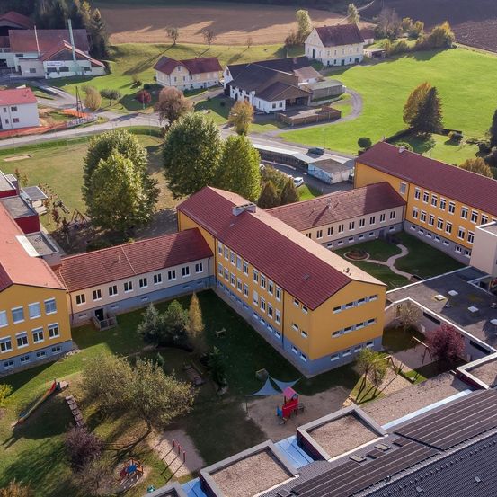 Ein Luftbild eines Schulgebäudes mit rotem Dach und gelben Wänden. Es hat mehrere Fenster und ist von Bäumen und einem kleinen Spielplatz umgeben.