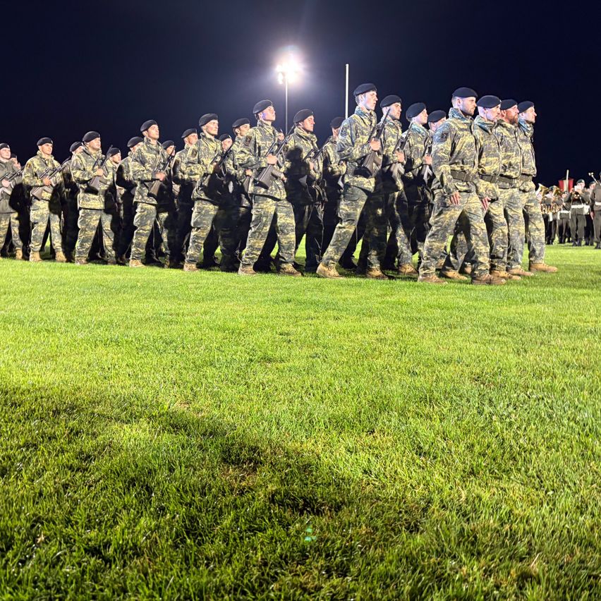 Eine Gruppe Soldaten in Camouflage-Uniformen, die Gewehre halten, marschiert nachts in Formation auf einem Rasenfeld.