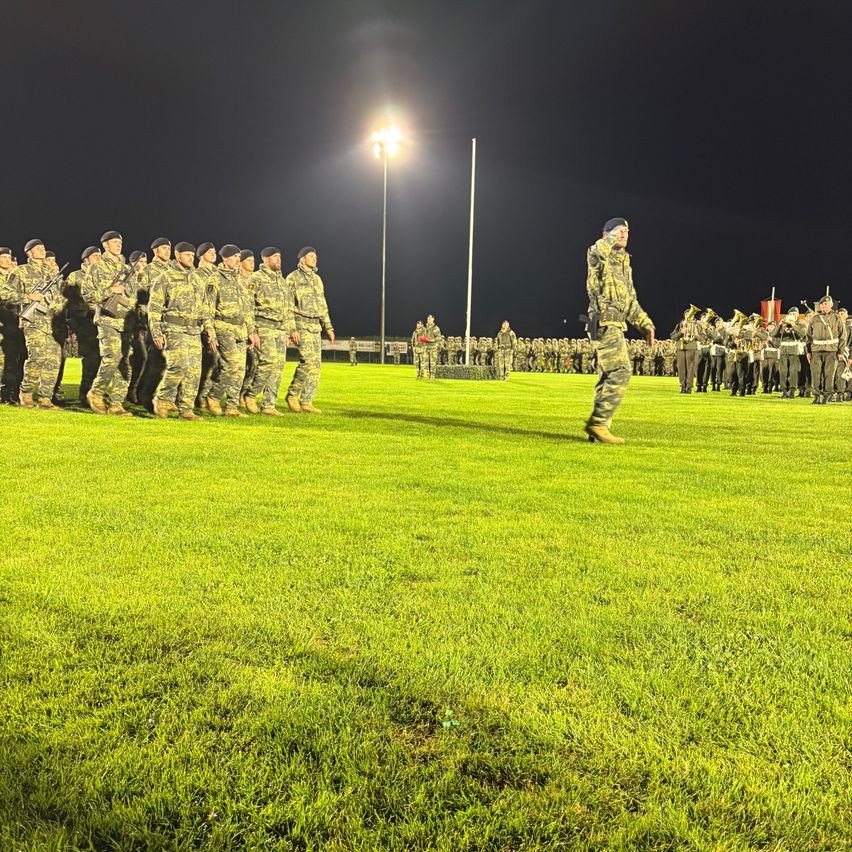 Eine Gruppe Soldaten in Tarnuniformen steht bei Nacht auf einem Feld. Ein Offizier geht vor ihnen. Dahinter steht eine Band mit Blechblasinstrumenten. Eine Straßenlaterne beleuchtet das Gebiet.