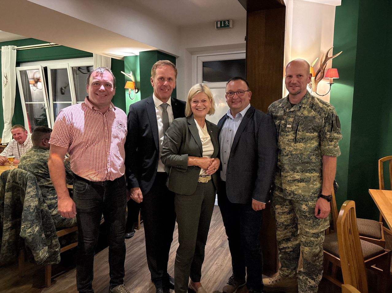Fünf Menschen posieren für ein Foto in einem Raum. Eine Frau in der Mitte lächelt und hält die Hand eines Mannes. Der Mann im Anzug schaut in die Kamera. Der Mann in der Militäruniform lächelt. Der Raum hat einen Holzboden und eine weiße Wand.