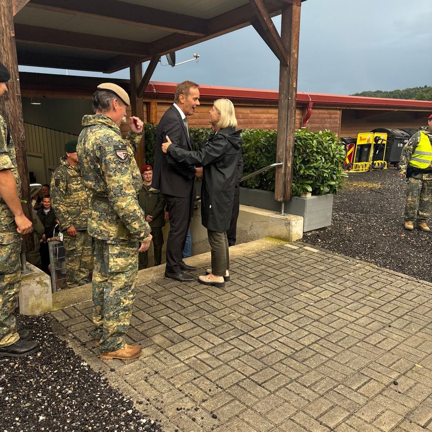 Mehrere Militärangehörige in Tarnuniformen stehen unter einem Vordach. Ein Mann in einem Anzug und eine Frau in einer Regenjacke führen ein Gespräch. Im Hintergrund befinden sich Pflanzen und Müllcontainer.