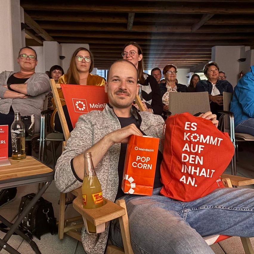 Ein Mann sitzt in einem Stuhl und hält eine Tasche, lächelt. Er hält eine Flasche und eine Tasche mit dem Text ES KOMMT AUF DEN INHALT AN. Hinter ihm sitzen Menschen auf Stühlen, darunter eine Frau mit Brille.