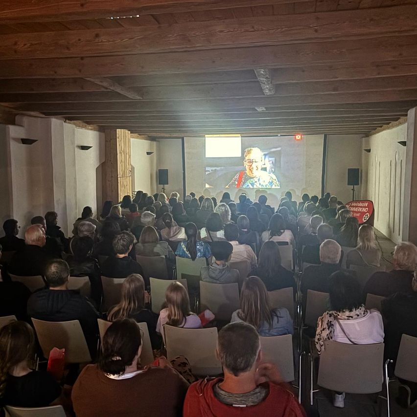 Eine Menschenmenge sitzt in Stühlen vor einer Projektionswand. Die Leinwand zeigt eine Frau mit Brille. Lautsprecher befinden sich auf beiden Seiten der Leinwand. Der Raum hat Holzbalken an der Decke.