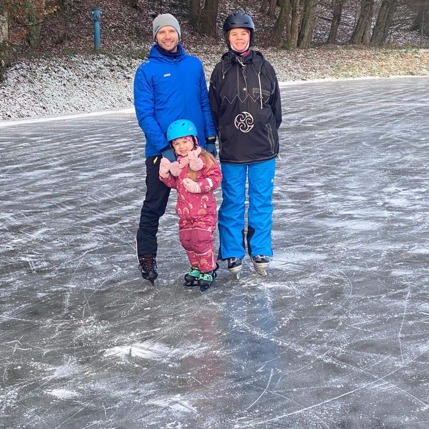 Drei Personen, zwei Erwachsene und ein Kind, skaten auf einer Eisbahn, wobei die Erwachsenen lächeln und das Kind einen Helm trägt.