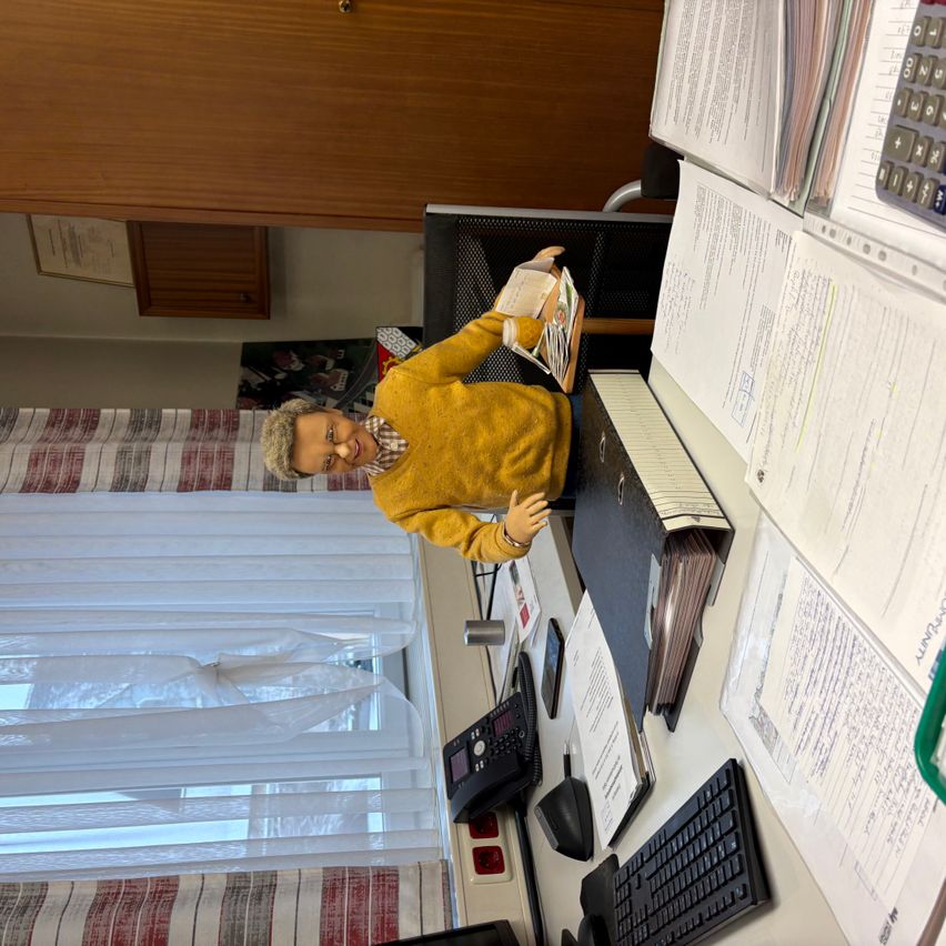 Eine Puppe in einem gelben Pullover steht an einem Schreibtisch in einem Büro. Papiere, ein Taschenrechner und ein Computer sind auf dem Schreibtisch. Das Büro hat ein Fenster mit weißen Vorhängen.