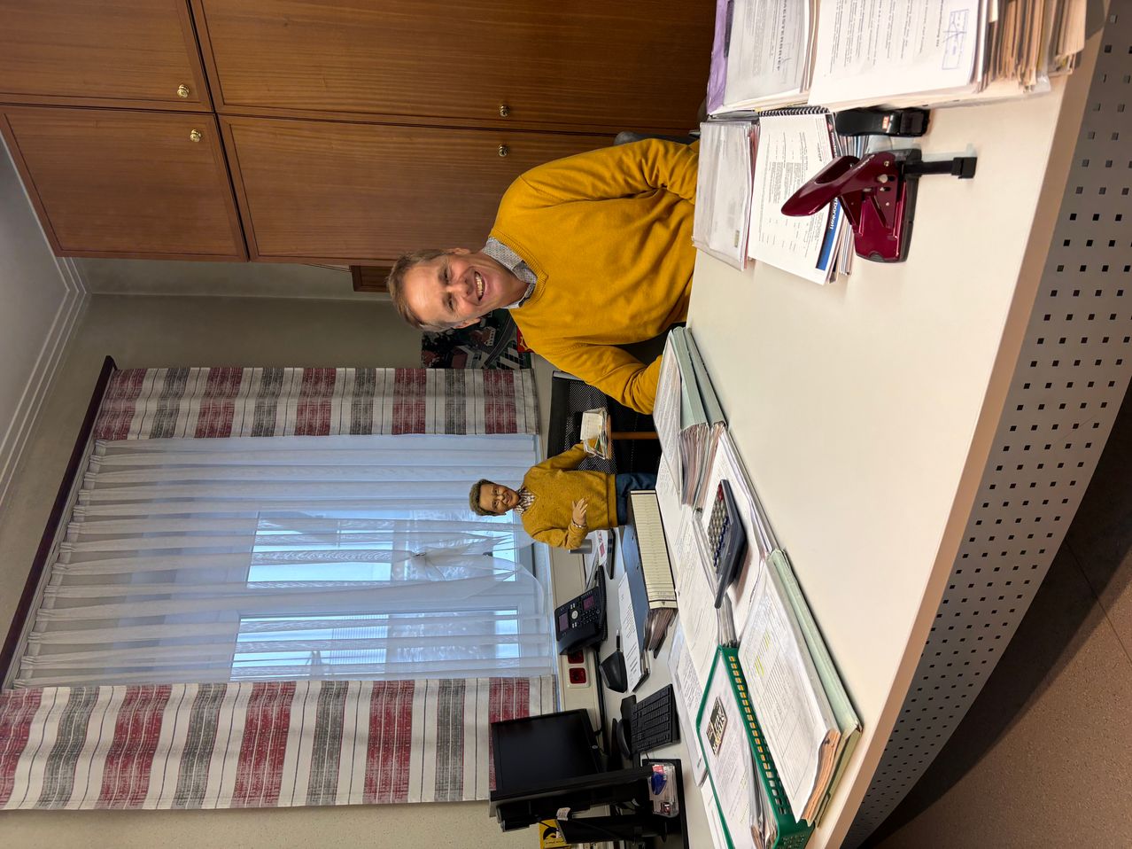 Ein Mann in einem gelben Pullover steht hinter einem Büroschreibtisch mit einer Mini-Puppe, einem Computer, einem Hefter und verschiedenen Dokumenten. Der Schreibtisch ist überladen, und im Hintergrund befindet sich ein Fenster mit Vorhängen.