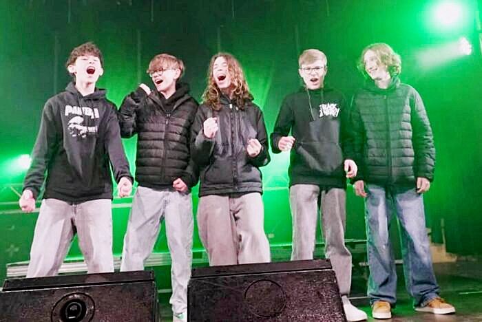 Fünf junge Männer, alle in dunklen Jacken gekleidet, singen auf einer Bühne. Sie haben den Mund offen und scheinen Spaß zu haben. Hinter ihnen ist eine grüne Wand mit Scheinwerfern.