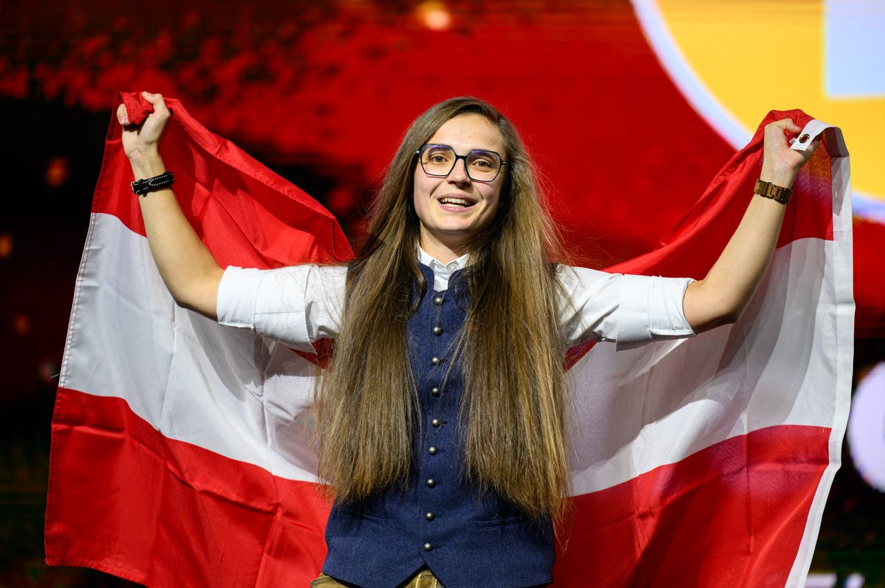 Eine lächelnde junge Frau mit langen Haaren hält eine rot-weiße Flagge, vermutlich bei einem Wettbewerb oder einer Veranstaltung. Sie trägt eine Brille und eine Weste mit Knöpfen. Der Hintergrund ist ein leuchtender rot-gelber Farbverlauf.