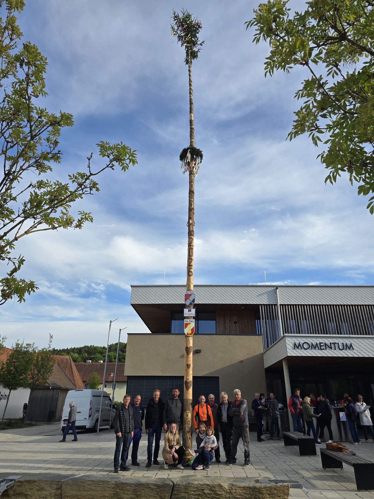 Eine Gruppe von Menschen steht um einen hohen Holzpfahl mit einer Fahne herum, vor einem Gebäude mit der Aufschrift 'Momentum'. Bäume und ein Van sind ebenfalls vorhanden.