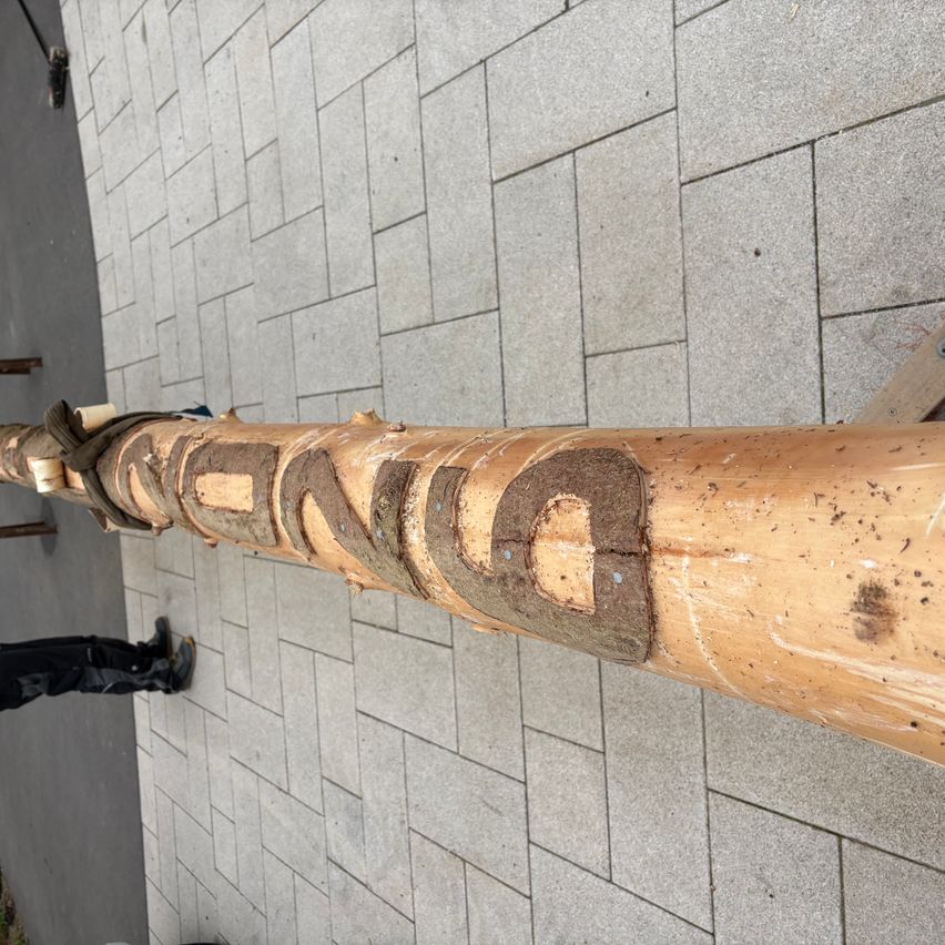Ein Nahaufnahme eines Holzpfahls mit dem eingravierten Wort 'tanz', auf einem gefliesten Boden platziert. Eine Person steht in der Nähe, gekleidet in Schwarz.