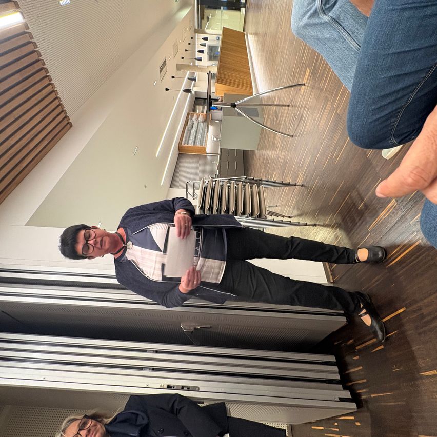 Ein Mann mit Brille hält ein Papier in der Hand und scheint mit jemandem zu sprechen. Dahinter sind Stühle gestapelt. Auf der rechten Seite sitzt eine Person, deren Hand sichtbar ist.