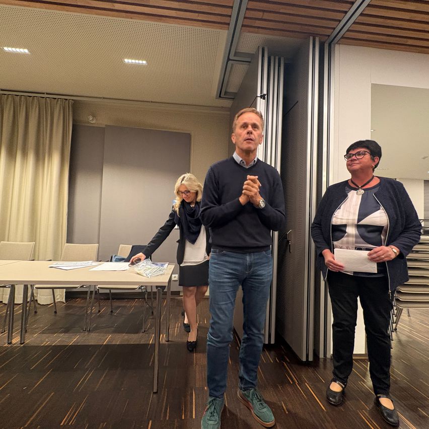 Drei Menschen stehen in einem Raum, ein Mann klatscht und zwei Frauen schauen auf Papiere. Ein Tisch mit Papieren und Stühlen steht hinter ihnen, mit Vorhängen und einer Tür an den Seiten.