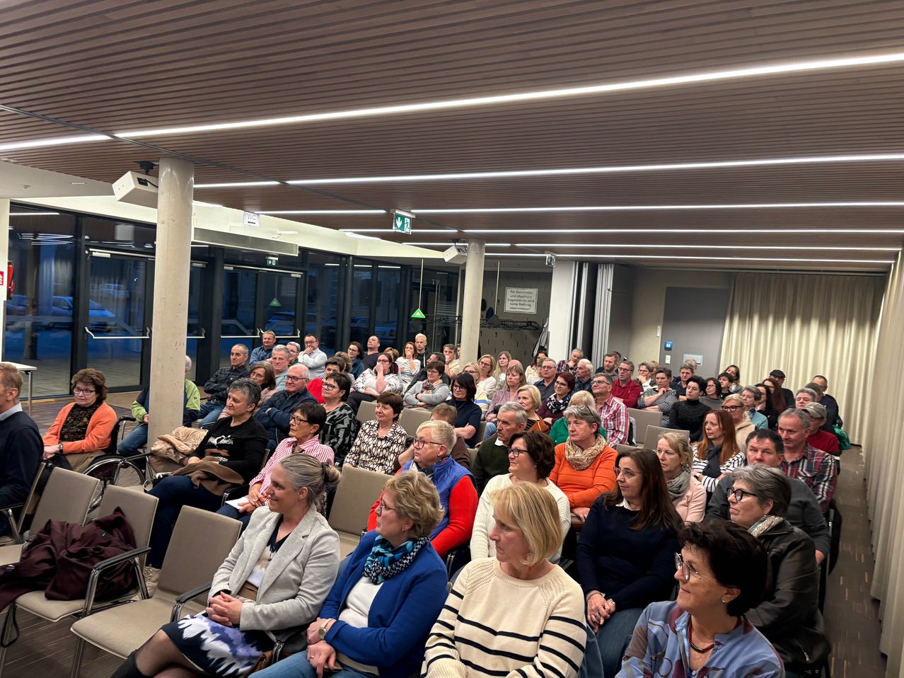 Eine große Gruppe von Menschen sitzt in einem überdachten Auditorium, einige mit Brillen und andere mit Schals. Der Raum ist gut beleuchtet, und die Decke hat Lampen und eine Überwachungskamera.