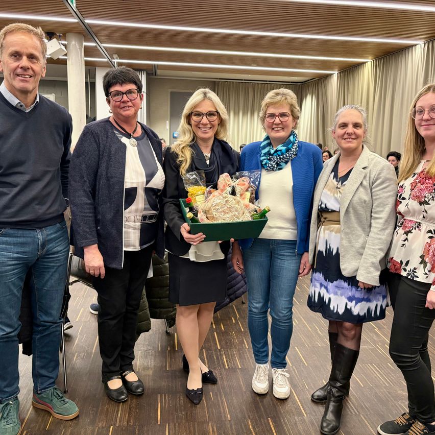 Eine Gruppe von Menschen steht in einem Raum und lächelt. Eine Frau hält einen Geschenkkorb. Hinter ihnen sind Vorhänge und Lichter zu sehen.