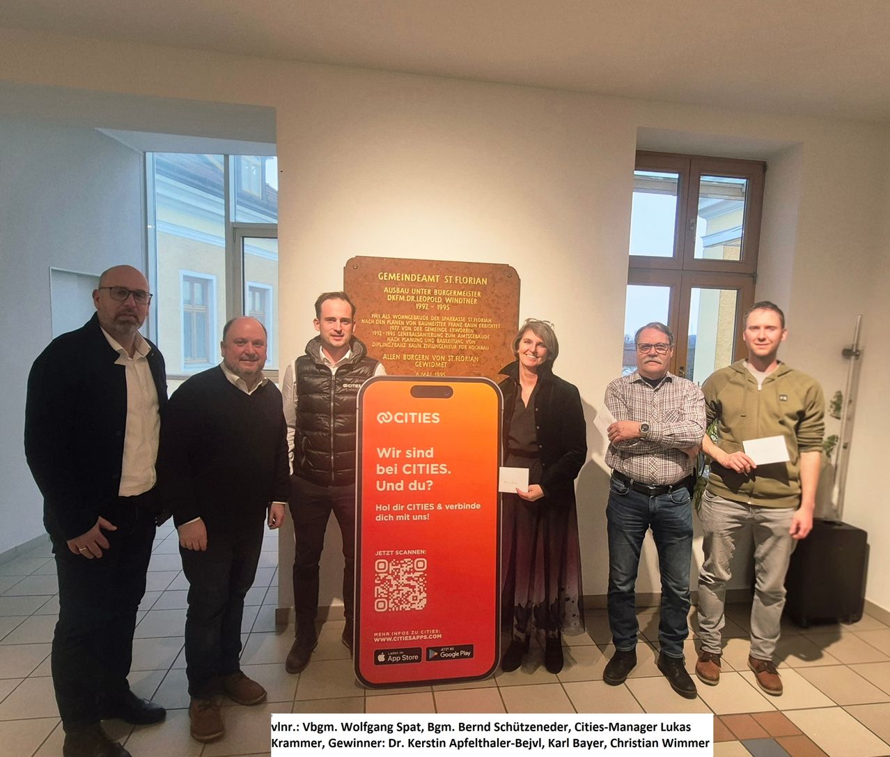 Fünf Personen stehen vor einer Plakette mit QR-Code und Smartphone. Sie lächeln und scheinen einen Erfolg zu feiern. Die Plakette enthält eine historische Erzählung, und die Gruppe besteht aus Männern und Frauen.