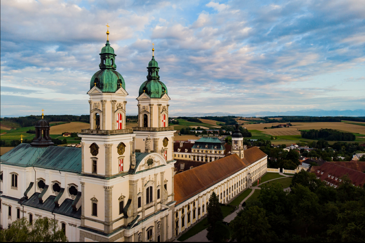 Luftaufnahme einer großen Kirche mit zwei grünen Kuppeln, rot-weiß karierten Flaggen und einem braunen Dach. Die Kirche ist von Bäumen und einer weiten Landschaft umgeben.
