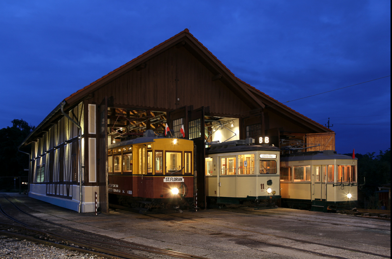 Zwei alte Straßenbahnen stehen nachts in einer Halle. Eine Straßenbahn hat ihre Scheinwerfer eingeschaltet, während die andere im Hintergrund geparkt ist. Die Halle hat ein rotes Dach und ist von Lichtern beleuchtet.