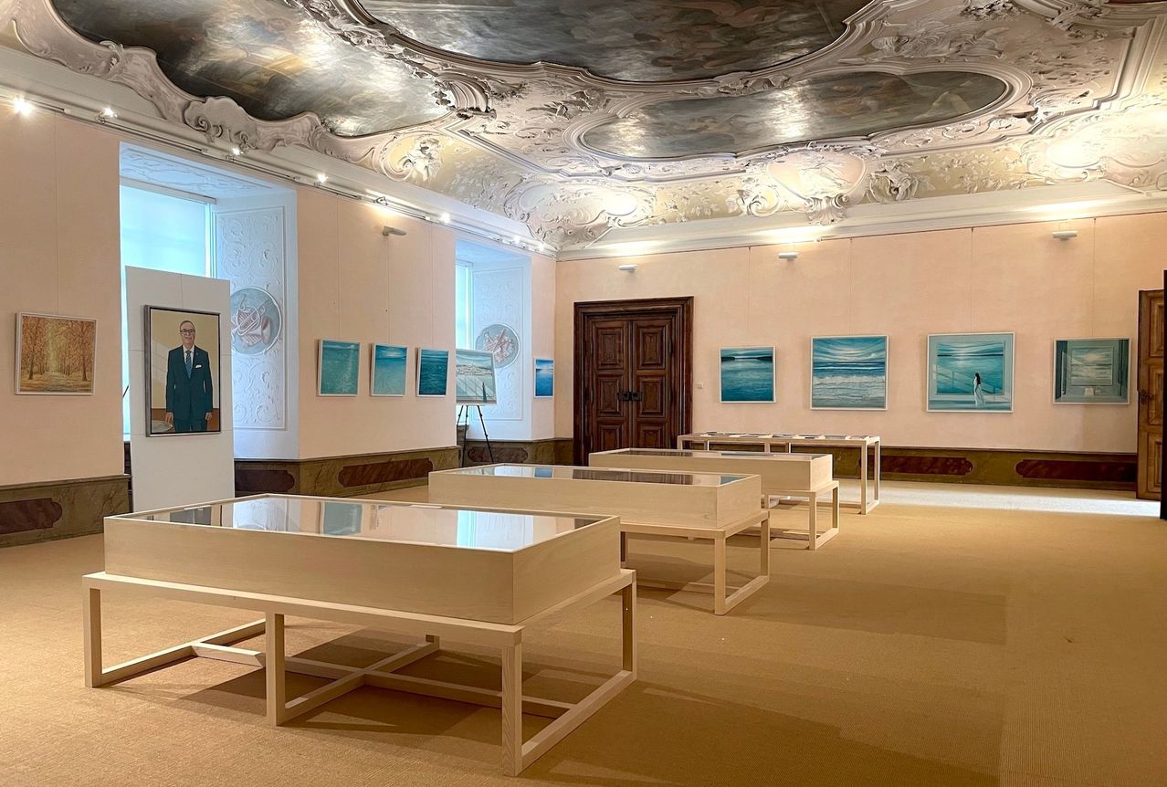 Eine Kunstgalerie mit einer verzierten Decke, einer Tür und mehreren Glasschaukästen. Kunstwerke schmücken die Wände, und ein Porträt eines Mannes befindet sich links.