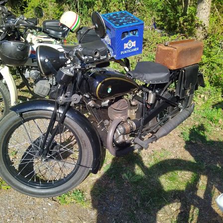 Ein schwarzes Oldtimer-Motorrad mit einer blauen Box mit der Aufschrift 'Puntigamer' und einer braunen Satteltasche steht auf dem Gras. Dahinter sind ein weiteres Motorrad und ein Helm zu sehen.