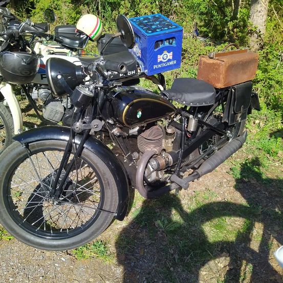 Ein schwarzes Oldtimer-Motorrad mit einer blauen Box mit der Aufschrift 'Puntigamer' und einer braunen Satteltasche steht auf dem Gras. Dahinter sind ein weiteres Motorrad und ein Helm zu sehen.