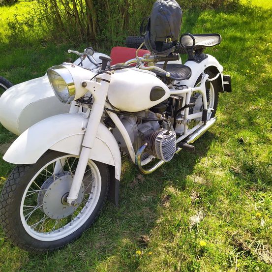 Ein weißes Vintage-Motorrad mit Seitenwagen und einer schwarzen Tasche auf dem Rücken steht auf einem Grasfeld.