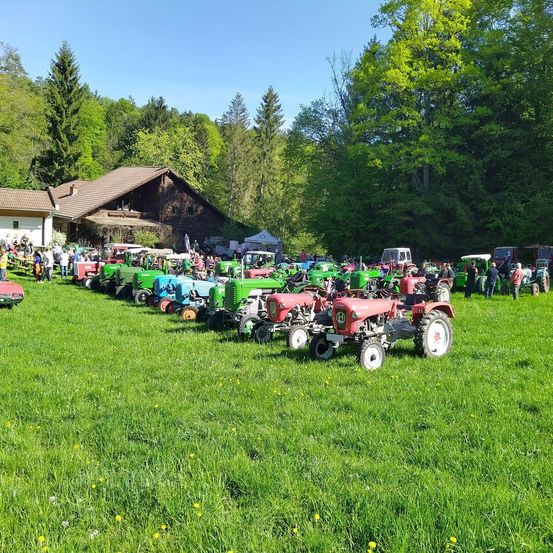 Eine Reihe von Traktoren in verschiedenen Farben ist auf einer Wiese geparkt, mit Menschen, die herumlaufen, und einem Gebäude im Hintergrund.