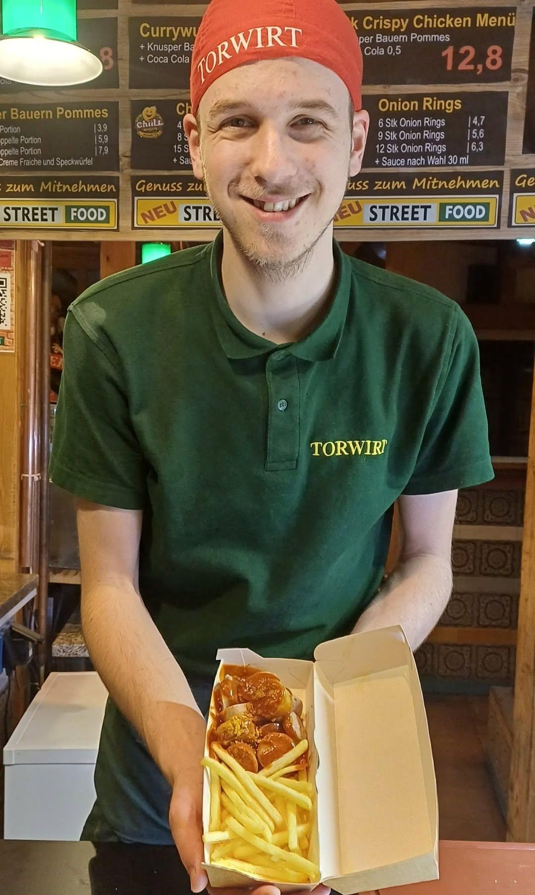 Ein lächelnder Mann mit einem grünen Poloshirt, auf dem 'TORWIR' steht, hält eine Box mit Straßenessen. Dahinter befindet sich ein Schild, das 'Street Food' anzeigt.