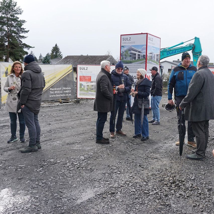 Eine Gruppe von Menschen versammelt sich auf einer Baustelle mit einem Baufahrzeug dahinter. Einige halten Getränke und unterhalten sich.